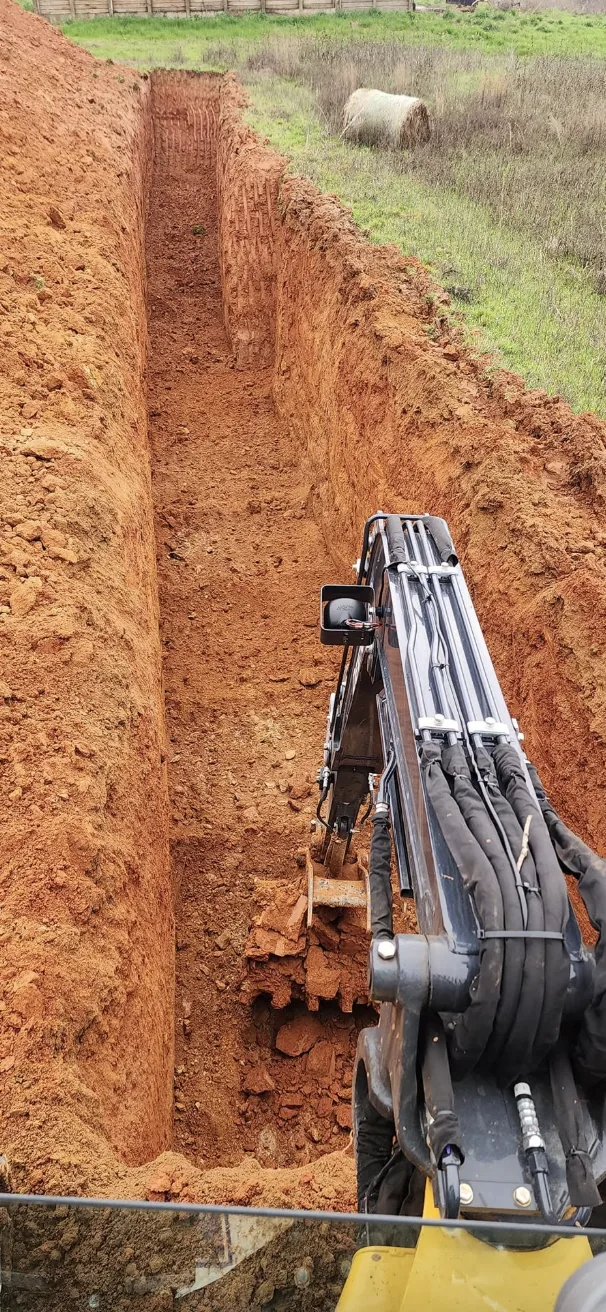 A deep trench cut through red clay with the excavator boom working in it &mdash; Little Acres Grading