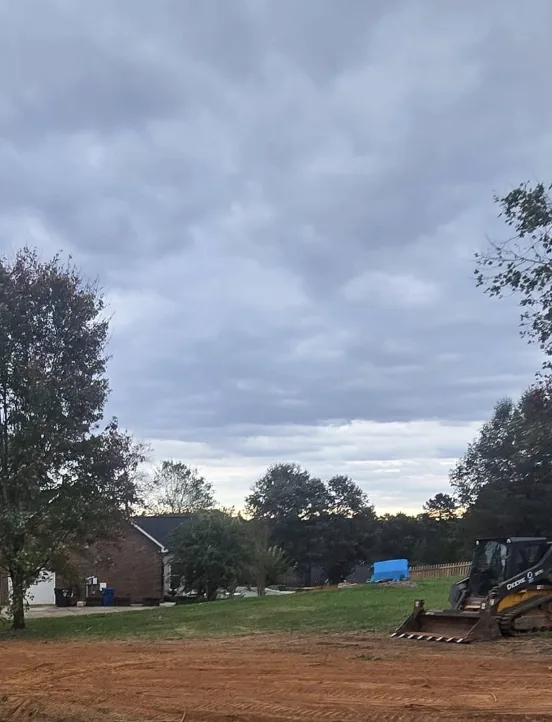 A skid steer finishing grade beside a brick home at dusk &mdash; Little Acres Grading