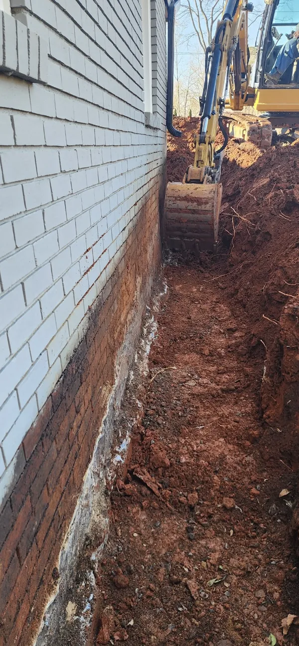 A foundation trench opened along the wall of a white brick home &mdash; Little Acres Grading